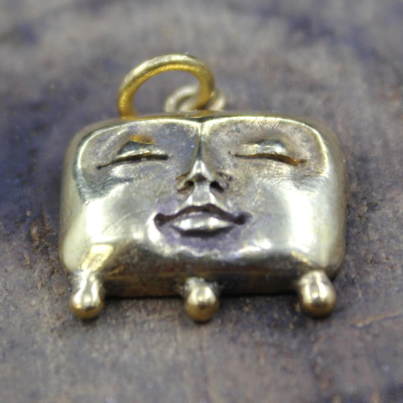 May include: A gold-colored pendant with a smiling face design. The pendant has a rectangular shape with rounded edges and small, ball-shaped feet. The face features closed eyes and a small, smiling mouth. A loop is at the top for hanging.