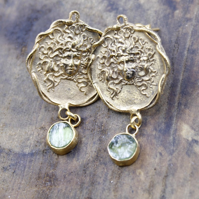 May include: A pair of gold-coloured earrings. Each earring has a circular pendant with a detailed face design and a dangling, round, light green stone. The earrings are displayed on a wooden surface.