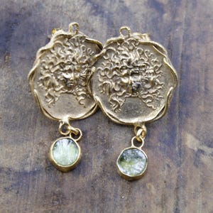 May include: A pair of gold-coloured earrings. Each earring has a circular pendant with a detailed face design and a dangling, round, light green stone. The earrings have a vintage or antique aesthetic.