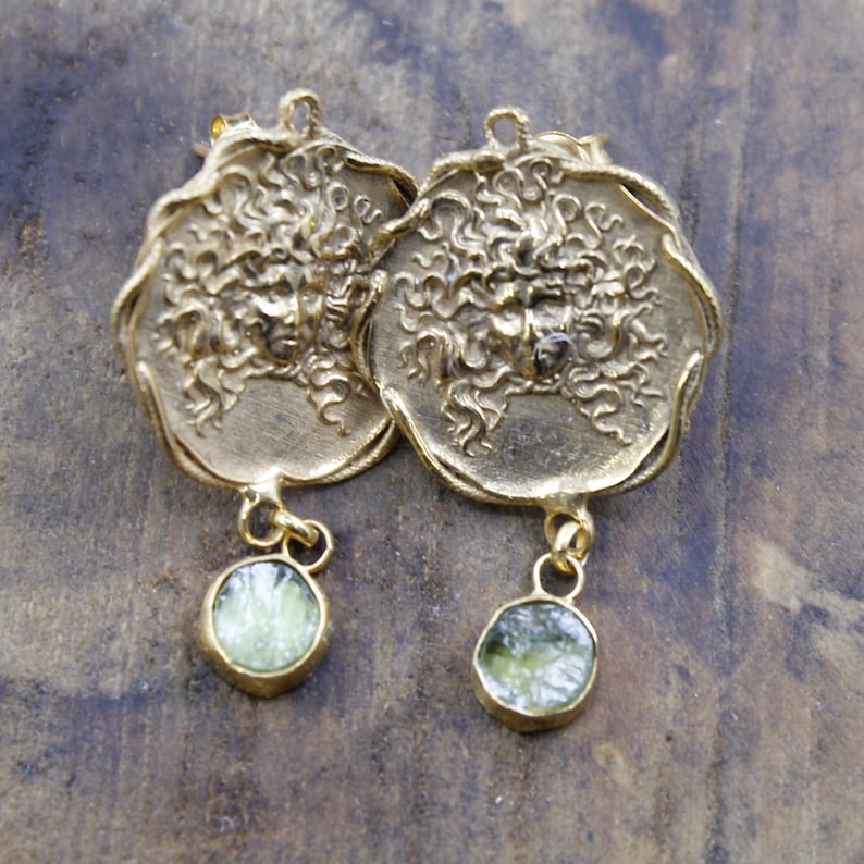 May include: A pair of gold-coloured earrings. Each earring has a circular pendant with a detailed face design and a dangling, round, light green stone. The earrings have a vintage or antique aesthetic.