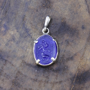 Puede incluir: Colgante ovalado azul cobalto con una fianza y bisel plateados. El colgante presenta un tallado en relieve detallado de una figura religiosa. El colgante está suspendido de un lazo plateado.