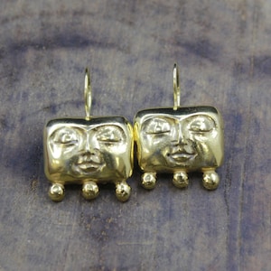 May include: A pair of gold-toned earrings featuring a rectangular face design with closed eyes and a smiling mouth. Each earring has three small spherical beads hanging from the bottom edge and a thin wire hook for attachment to the ear.