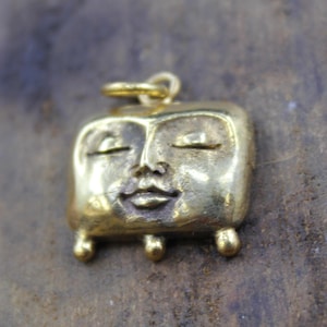 May include: A gold-colored pendant with a stylized face. The face has closed eyes, a small nose, and a smiling mouth. The pendant is square with rounded edges and small, ball-shaped feet. A loop is at the top for a chain.