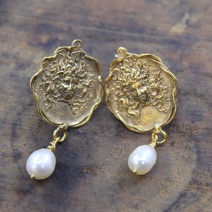 May include: A pair of gold-toned earrings. Each earring features a detailed face design with flowing hair, and a dangling white pearl. The earrings have a vintage or antique aesthetic.