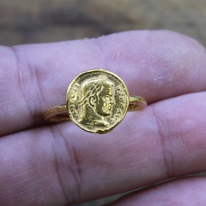 May include: A gold ring featuring a circular coin-style centerpiece. The face of the coin displays a relief profile of a figure wearing a laurel wreath, surrounded by faint, embossed lettering along the edge of the circular gold metal surface.