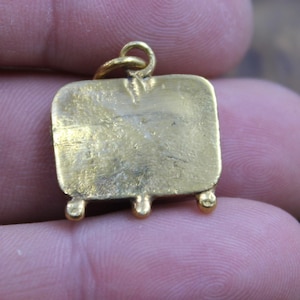 May include: A small, gold-colored pendant with a rectangular body and three small, rounded feet. The pendant has a loop at the top for a chain. The surface shows slight wear.