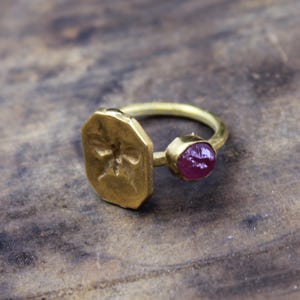 May include: A gold-colored ring with two distinct features. One side has a geometric, engraved design, and the other features a round, deep red gemstone. The ring is set against a textured, brown surface.