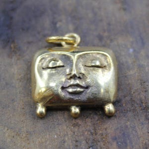 Puede incluir: Un colgante dorado con un diseño de rostro sereno. El colgante tiene forma cuadrada con esquinas redondeadas, ojos cerrados y una boca pequeña. Cuenta con tres pequeños pies redondeados y un lazo para una cadena.