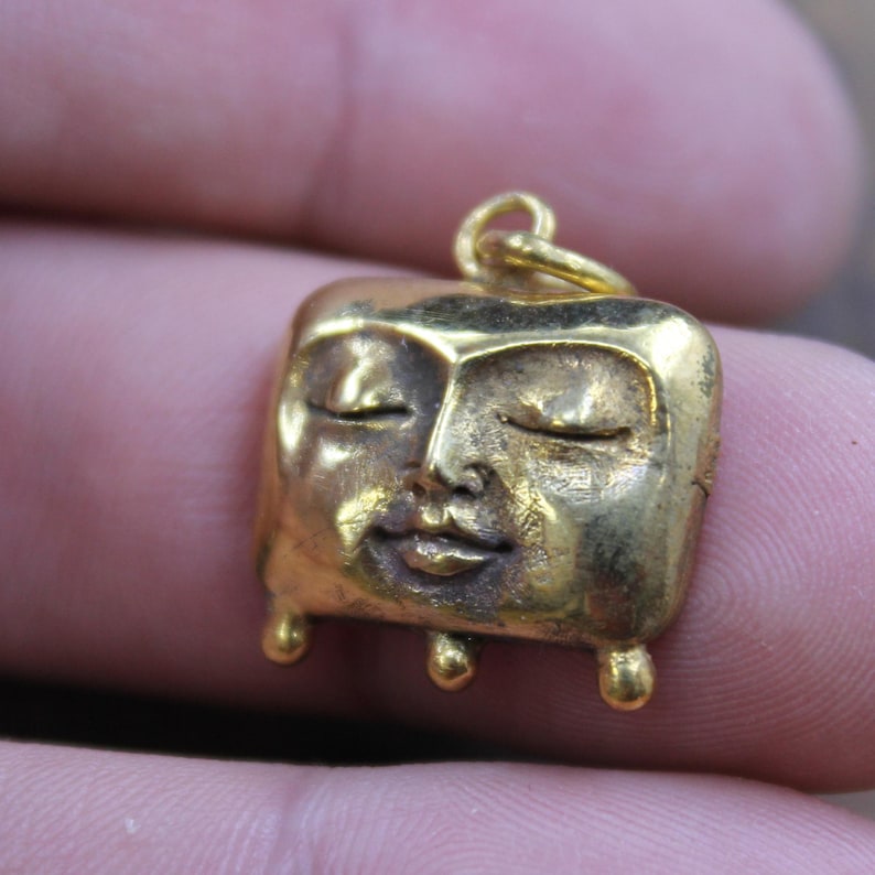 May include: A small, gold-colored pendant featuring a stylized face with closed eyes. The pendant has a square shape with rounded edges and three small, spherical feet. The bail is a simple loop for attaching to a chain or cord.