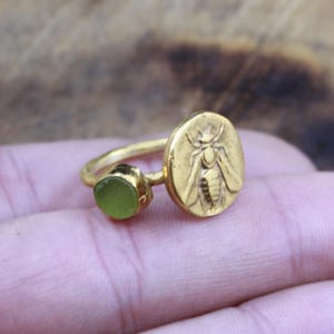 May include: A gold-colored ring featuring a circular design with a bee engraving and a small, round, light green stone. The ring has a delicate band and a vintage aesthetic. The bee design is detailed and prominent.