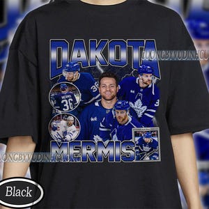 May include: Black t-shirt featuring a collage of hockey players in blue and white uniforms. The shirt prominently displays the name "DAKOTA MERMIS" in a bold, blue, and white font. Additional text and player images are incorporated into the design.