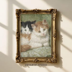 May include: A framed vintage-style painting featuring two kittens. One kitten is black and white, the other is orange and white. The painting is in a gold ornate frame. The text "DEVÁZYN STUDIO" is at the bottom of the frame.