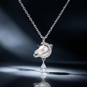 May include: A silver-toned necklace featuring a pendant shaped like a planet with a central white pearl, surrounded by a ring encrusted with small clear stones. A single teardrop-shaped clear stone hangs from the bottom of the planet pendant.