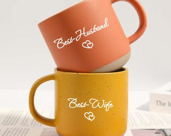 May include: Two ceramic mugs, one orange and one yellow, stacked together. The orange mug has the text "Best Husband" and two hearts, while the yellow mug says "Best Wife" with two hearts. Both mugs have a speckled texture.