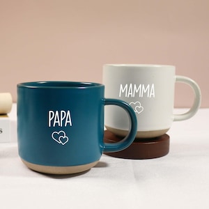 Puede incluir: Dos tazas de cerámica. Una es de color verde azulado con "PAPA" y dos corazones blancos. La otra es blanca con "MAMMA" y dos corazones blancos. Ambas tazas tienen una base beige y un asa curva.
