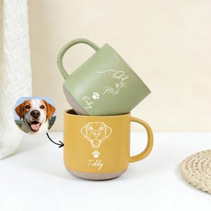 Può includere: Due tazze in ceramica, una verde e una gialla, impilate l'una sull'altra. La tazza verde ha un'illustrazione di gatto e il nome "Ruby". La tazza gialla presenta un'illustrazione di cane e il nome "Teddy". Entrambe le tazze hanno impronte di zampe.