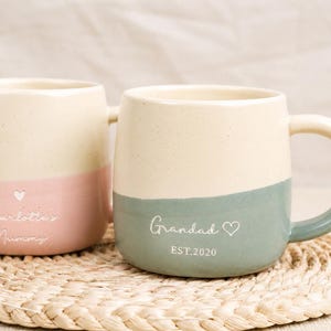 Peut inclure: Deux mugs en céramique bicolores. L'un est rose et crème, avec le texte "Charlotte's Mummy". L'autre est vert et crème, avec le texte "Grandad EST.2020". Les deux mugs ont un symbole de cœur.