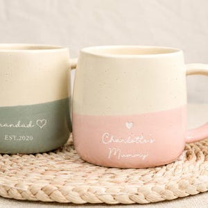 Op de afbeelding: Twee keramische mokken met een crème- en pastelkleurig blokontwerp. Eén mok is saliegroen en zegt "Grandad" met een hart en "EST.2020". De andere is roze en zegt "Charlotte's Mummy" met een hart.