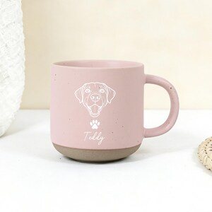 Puede incluir: Una taza de cer&aacute;mica rosa con base y asa marr&oacute;n. La taza presenta una ilustraci&oacute;n blanca de la cara de un perro, una huella de pata y el nombre "Teddy". La taza est&aacute; sobre una superficie blanca.