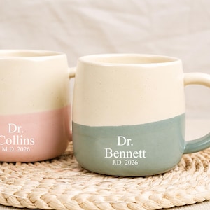 Könnte beinhalten: Zwei Keramikbecher mit einem gesprenkelten cremefarbenen oberen Teil und einem einfarbigen unteren Teil. Der linke Becher ist rosa mit weißem Text Dr. Collins M.D. 2026. Der rechte Becher ist salbeigrün mit weißem Text Dr. Bennett J.D. 2026.