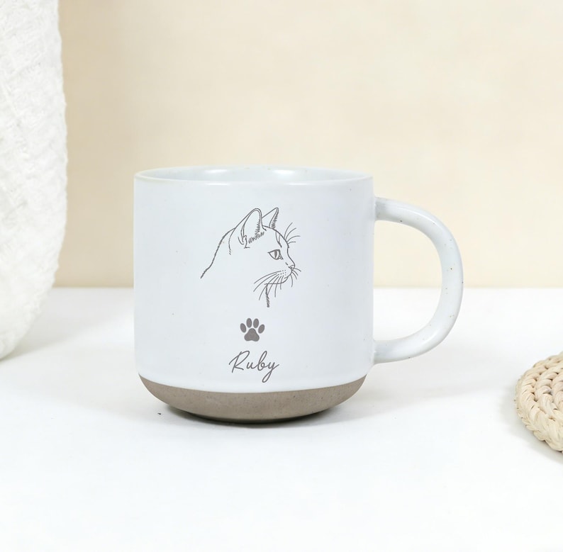Puede incluir: Taza de cer&aacute;mica blanca con base y asa grises. La taza presenta un dibujo lineal negro del perfil de un gato, una huella de pata y el nombre "Ruby" en cursiva. La taza est&aacute; sobre una superficie blanca.