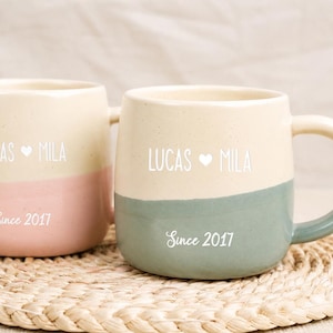 Pode incluir: Duas canecas de cerâmica com a parte superior creme e a inferior colorida. Uma caneca tem a parte inferior rosa, a outra verde. Cada caneca tem os nomes "LUCAS ♥ MILA" e "Since 2017". As canecas estão sobre uma esteira tecida.
