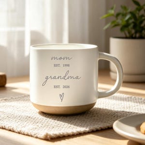 Puede incluir: Una taza de cerámica blanca con base marrón, con las palabras "mom" y "grandma" en escritura gris, junto con los años "EST. 1998" y "EST. 2026". Un pequeño símbolo de corazón está debajo de la palabra "grandma". La taza está sobre un mantel individual de color neutro.