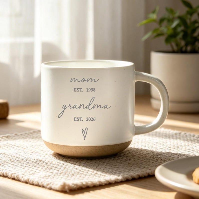 Puede incluir: Una taza de cer&aacute;mica blanca con base marr&oacute;n, con las palabras "mom" y "grandma" en escritura gris, junto con los a&ntilde;os "EST. 1998" y "EST. 2026". Un peque&ntilde;o s&iacute;mbolo de coraz&oacute;n est&aacute; debajo de la palabra "grandma". La taza est&aacute; sobre un mantel individual de color neutro.