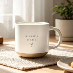 Puede incluir: Taza de cerámica color crema con el texto "alden's mama" y un símbolo de corazón. La base de la taza es de color marrón claro. La taza está sobre un salvamanteles beige.
