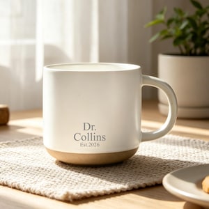 Könnte beinhalten: Ein cremefarbener Keramikbecher mit glatter Oberfläche und einem naturfarbenen braunen Boden. Auf der Vorderseite des Bechers befindet sich der graue Aufdruck Dr. Collins und Est. 2026. Der Becher hat einen geschwungenen Henkel und eine zylindrische Form.