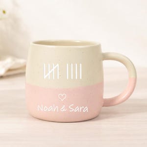 May include: A two-tone ceramic mug with a cream upper half and a pink lower half. The mug features a tally mark design and the names "Noah & Sara" with a heart symbol. The handle is also pink.