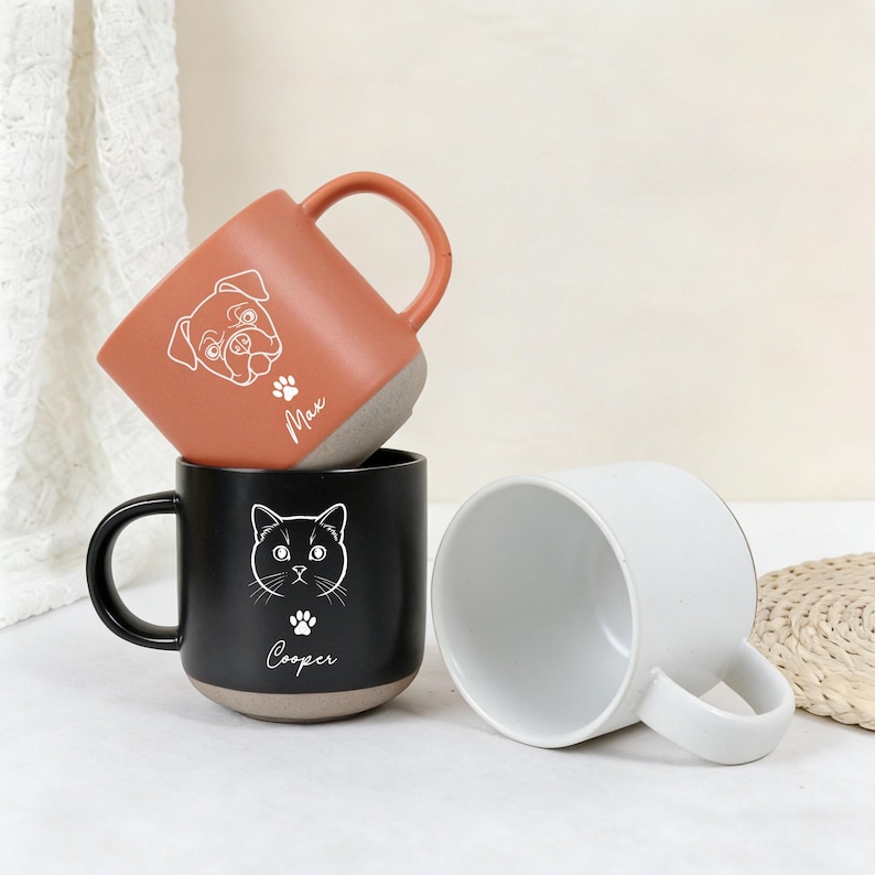 Puede incluir: Tres tazas de cer&aacute;mica con dise&ntilde;os tem&aacute;ticos de mascotas. Una es de color naranja quemado con una ilustraci&oacute;n de un pug y el nombre "Max". Otra es negra con una ilustraci&oacute;n de gato y el nombre "Cooper". La tercera taza es blanca.
