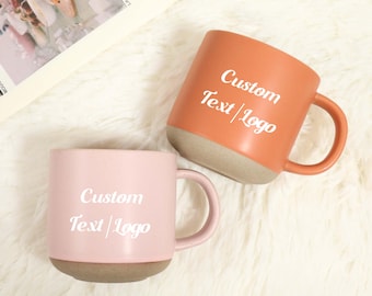 Op de afbeelding: Twee keramische mokken, één perzik en één roze, met een neutrale basis en handvat. Elke mok heeft de woorden "Custom Text/Logo" in het wit gedrukt. De mokken staan op een zacht, wit oppervlak.