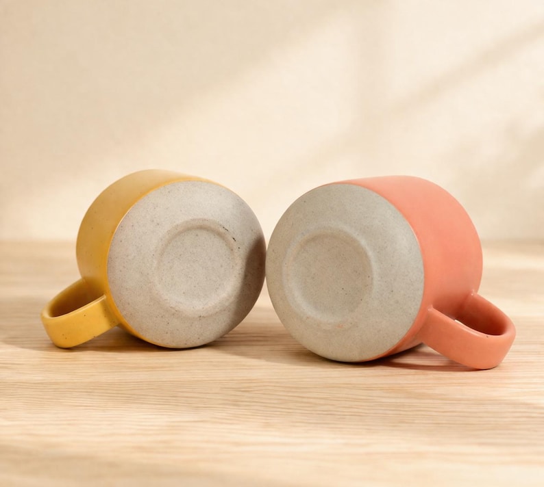 Puede incluir: Dos tazas de cer&aacute;mica, una amarilla y otra coral, se muestran sobre una superficie de madera. Las tazas est&aacute;n posicionadas con sus bases hacia arriba, revelando una base texturizada de color gris claro. Cada taza tiene un asa redondeada.