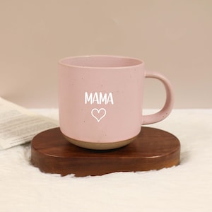 Pode incluir: Uma caneca de cerâmica rosa com a palavra "MAMA" e um símbolo de coração impresso em branco. A caneca tem uma textura salpicada e uma base marrom, colocada em uma base de madeira.