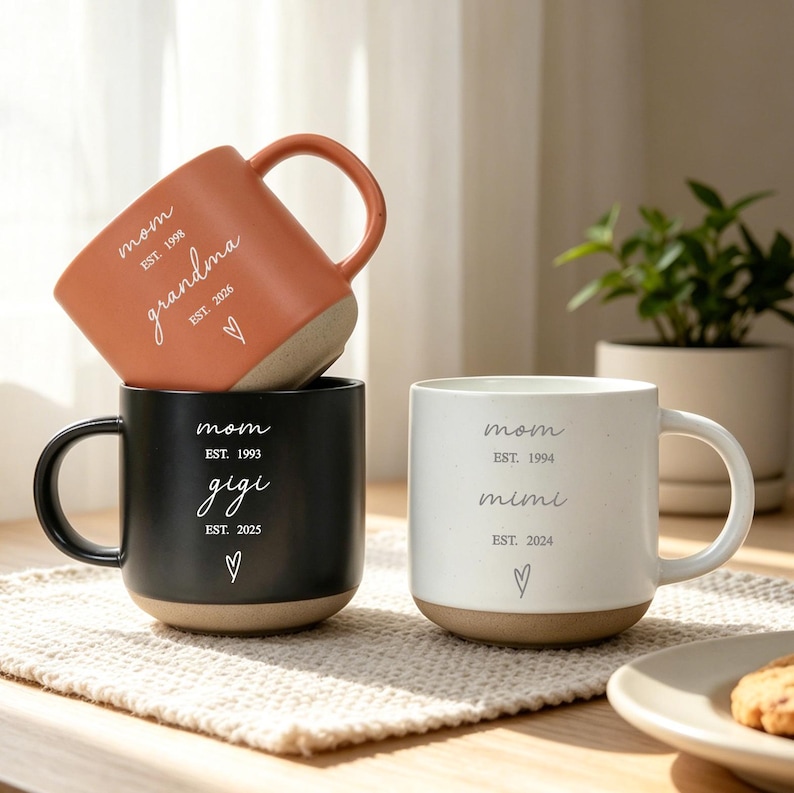 Puede incluir: Tres tazas de cer&aacute;mica en tonos terracota, negro y blanco. Cada taza presenta la palabra "mam&aacute;" y un nombre diferente, como "abuela", "gigi" o "mimi", junto con los a&ntilde;os de establecimiento y un s&iacute;mbolo de coraz&oacute;n.