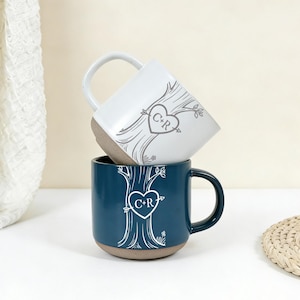 Peut inclure: Deux mugs en céramique, un blanc et un bleu canard, empilés. Chaque mug présente un motif d'arbre avec un emblème en forme de cœur avec les initiales "C+R". Les mugs ont une base marron et une anse blanche.