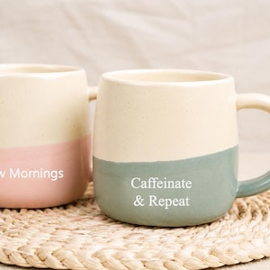 Può includere: Due tazze in ceramica con la parte superiore color crema e la parte inferiore colorata. Una tazza ha la base rosa con il testo "Slow Mornings". L'altra ha la base verde con il testo "Caffeinate & Repeat". Entrambe le tazze sono su un sottobicchiere intrecciato.