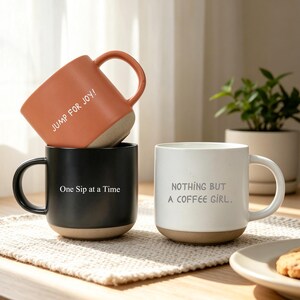 Op de afbeelding: Drie keramische mokken met oren en ongeglazuurde onderkanten. Een terracotta mok heeft de tekst JUMP FOR JOY!, een zwarte mok heeft de tekst One Sip at a Time en een witte gespikkelde mok heeft de tekst NOTHING BUT A COFFEE GIRL.