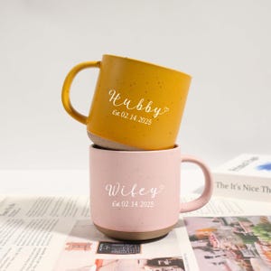 Puede incluir: Dos tazas de cerámica, una amarilla mostaza y otra rosa, apiladas una encima de la otra. La taza amarilla dice "Hubby" con la fecha "Est. 02.14.2025". La taza rosa dice "Wifey" con la misma fecha. Ambas tazas tienen una textura moteada.