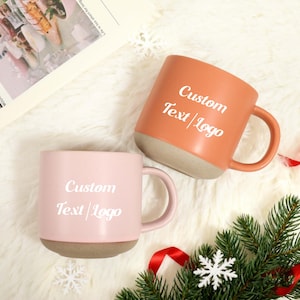 Könnte beinhalten: Zwei Keramikbecher, einer pfirsichfarben und einer rosa, mit dem Text "Custom Text/Logo" in Weiß. Beide Becher haben einen beigefarbenen Boden und passende Henkel. Die Becher stehen auf einer weißen, flauschigen Oberfläche, mit einem Tannenzweig und einer Schneeflocke.