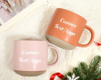Puede incluir: Dos tazas de cerámica, una melocotón y otra rosa, con el texto "Custom Text/Logo" en blanco. Ambas tazas tienen una base beige y asas a juego. Las tazas están sobre una superficie blanca y esponjosa, con una ramita de hoja perenne y un copo de nieve.