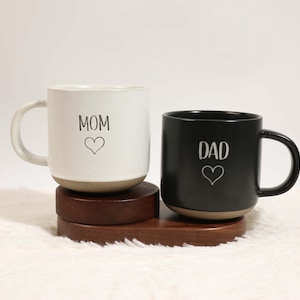 Puede incluir: Dos tazas de cerámica, una blanca y otra negra, sobre una bandeja de madera. La taza blanca tiene "MOM" y un corazón, la taza negra tiene "DAD" y un corazón. Ambas tazas tienen una base beige y un asa curvada.