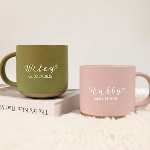 Op de afbeelding: Twee keramische mokken, één olijfgroen en één roze, met de woorden "Wifey" en "Hubby" in script, en de datum "Est. 02.14.2025". De mokken staan op een wit oppervlak.