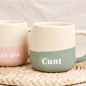 May include: Two ceramic mugs with a two-tone design. One mug is cream and pink with the text "FUCK OFF" in white. The other mug is cream and teal with the text "Cunt" in white. Both mugs have a rounded shape and a handle.