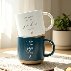 Puede incluir: Dos tazas de cer&aacute;mica apiladas. La taza superior es blanca con las palabras "mam&aacute;" y "abuela" impresas. La taza inferior es azul oscuro con las palabras "mam&aacute;" y "gigi" impresas. Ambas tazas tienen un s&iacute;mbolo de coraz&oacute;n.