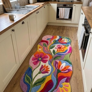 Könnte beinhalten: Ein unregelmäßig geformter Teppich mit einem lebendigen, mehrfarbigen Blumenmuster. Das Design zeigt stilisierte Blüten in den Farben Pink, Orange, Gelb, Lila und Blau auf einem cremefarbenen Hintergrund mit wellenförmigen, fließenden Linien.