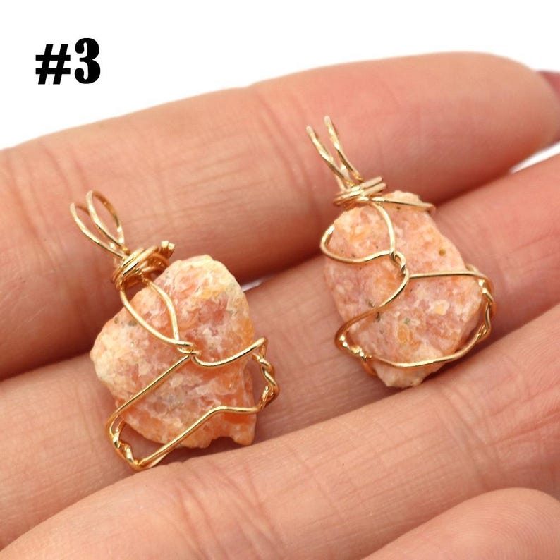 May include: Two raw, peach-colored stone pendants wrapped in gold-colored wire. The pendants are held in a hand. The number "#3" is in the upper left corner.