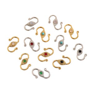 May include: A collection of decorative jewelry clasps in gold and silver tones. Each clasp features a central, oval-shaped gemstone in various colors, including red, green, blue, and pink. The clasps are designed with a curved, S-shaped hook.