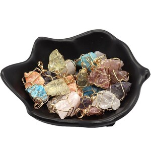 May include: A collection of colorful raw crystal pendants in a black bowl. The crystals are various colors, including pink, blue, yellow, and purple, and are wrapped in gold-colored wire. The bowl has a unique, organic shape.